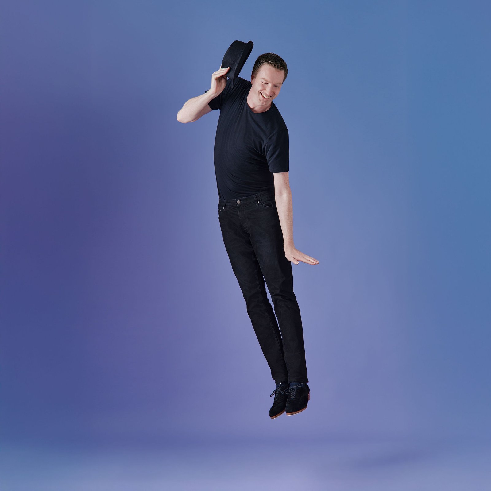 man in dark clothing jumps in the air, holding a hat in his raised right hand, against a blue and purple background
